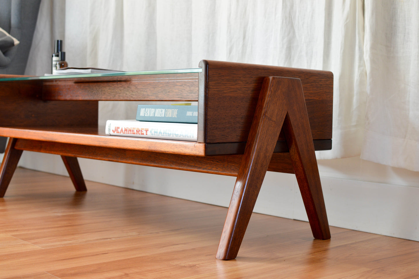  Mid Century Modern style coffee table with a clear glass top and brown wooden legs with a compass leg design. Magazines and books are placed on the table, adding to its aesthetic as a functional and stylish piece of furniture.